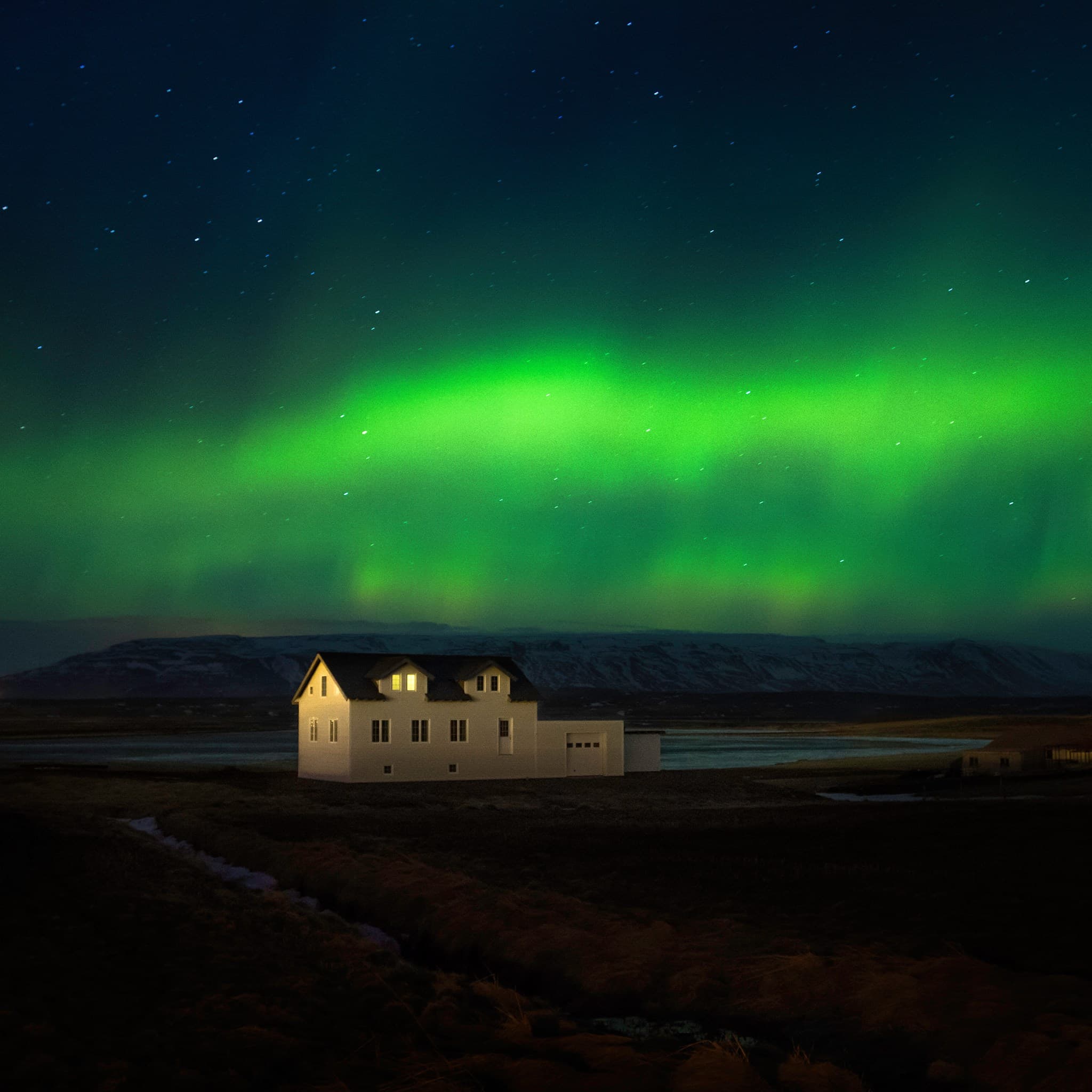 Mynd af Áshildarholti undir norðurljósum. Það er ljós í einum glugga og það er snjór á jörðu.